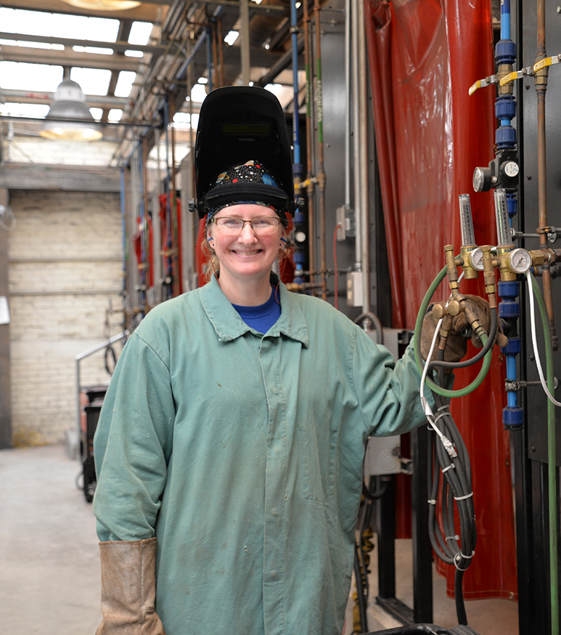 Female welder standing in SVHEC's welding lab. Her welding helmet is raised and she smiles directly at the camera.