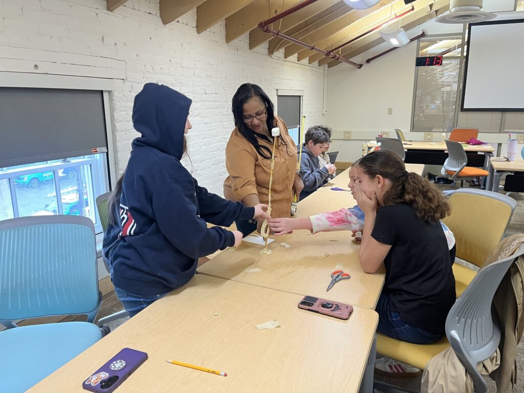 Entrepreneurship Education Coordinator Jocelyn Forest leads a session on the foundations of entrepreneurship to a Halifax County Homeschool Collaborative. In the photo students are building a tower made of spaghetti noodles with a marshmallow on top. Jocelyn and several students have their hands on  the spaghetti tower.
